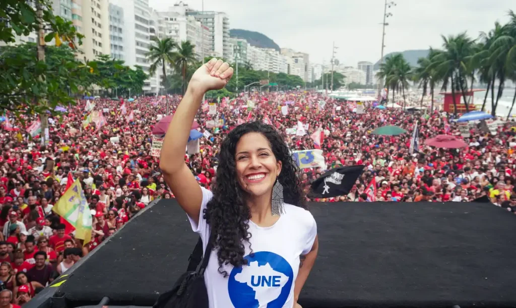 Uma mulher com camiseta da UNE ergue o punho em frente a multidão em manifestação ao ar livre