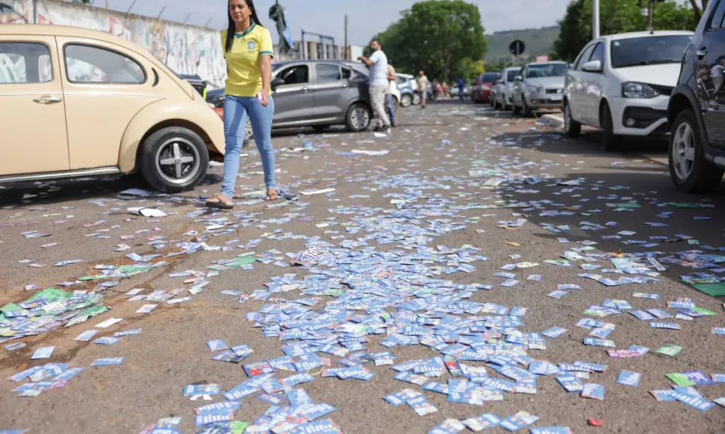 Uma mulher caminha por uma rua com papeladas espalhadas no chão e carros estacionados ao fundo