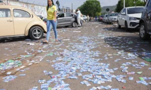 Uma mulher caminha por uma rua com papeladas espalhadas no chão e carros estacionados ao fundo