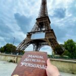 Mão segura livro intitulado "Manual Secreto da Persuasão" com a Torre Eiffel ao fundo