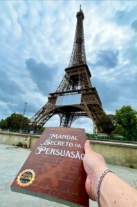Mão segura livro intitulado "Manual Secreto da Persuasão" com a Torre Eiffel ao fundo